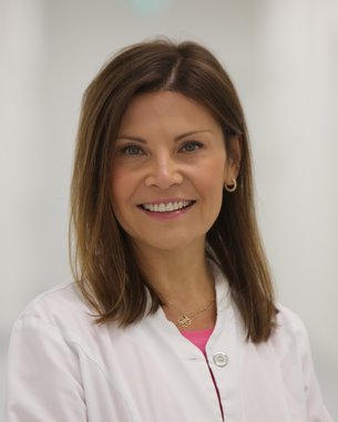 Eine lächelnde Frau in einem weißen Kittel steht in einem hellen, modernen Flur. Sie hat lange, braune Haare und trägt goldene Ohrringe.