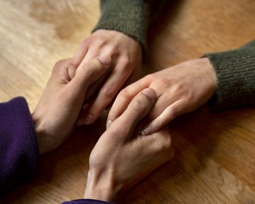 Zwei Hände halten sich sanft, während eine dritte Hand darüber gelegt ist. Der Fokus liegt auf der Berührung und Verbindung zwischen den Händen.