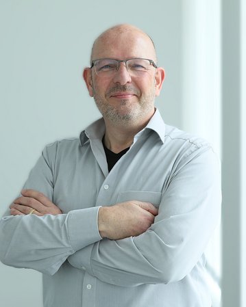 Ein Mann mit Brille und einem grauen Hemd steht mit verschränkten Armen vor einer hellen Wand.