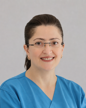 Eine Frau mit Brille trägt ein blaues medizinisches Outfit und lächelt freundlich in die Kamera. Der Hintergrund ist neutral und hell.