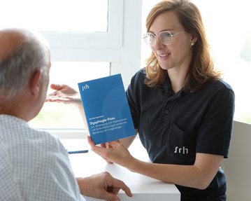 Eine Fachkraft zeigt einem Patienten ein Informationsheft über diagnostische Prozesse, während sie in einem hellen Raum an einem Tisch sitzen. Der Patient trägt ein Krankenhaushemd und hört aufmerksam zu.