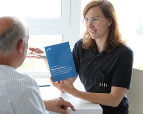 Eine Fachkraft zeigt einem Patienten ein Informationsheft über diagnostische Prozesse, während sie in einem hellen Raum an einem Tisch sitzen. Der Patient trägt ein Krankenhaushemd und hört aufmerksam zu.