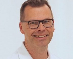 Ein Mann mit Brille und einem weißen Hemd lächelt freundlich in die Kamera. Der Hintergrund ist hell und neutral.