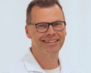 Ein Mann mit Brille und einem weißen Hemd lächelt freundlich in die Kamera. Der Hintergrund ist hell und neutral.