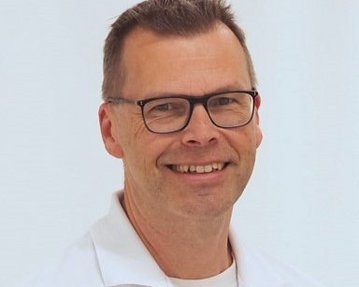 Ein Mann mit Brille und einem weißen Hemd lächelt freundlich in die Kamera. Der Hintergrund ist hell und neutral.