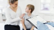 Eine Mutter sitzt neben ihrem Kind, während ein Arzt mit Handschuhen ein Formular ausfüllt. Das Kind erhält eine Impfung.