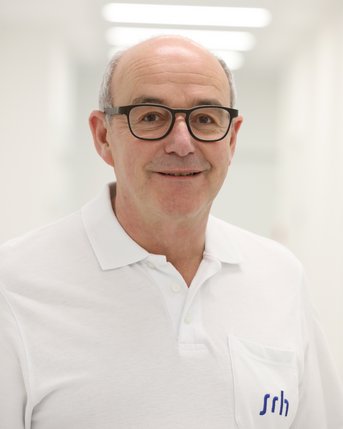 Ein Mann mit Brille trägt ein weißes Poloshirt mit dem Logo "srh" und lächelt freundlich in einem hellen, modernen Raum.