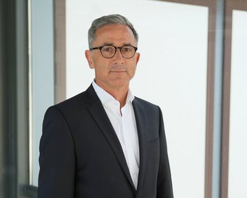 Ein Mann in einem Anzug steht vor einer Glaswand und schaut direkt in die Kamera. Er hat graue Haare und trägt eine Brille.