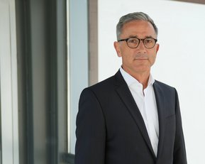 Ein Mann in einem Anzug steht vor einer Glaswand und schaut direkt in die Kamera. Er hat graue Haare und trägt eine Brille.