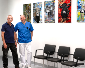 Zwei Männer stehen in einem modernen Raum mit Stühlen. An der Wand hängen farbenfrohe Tierporträts. Der Mann links trägt ein schwarzes Hemd, der rechts einen blauen Arztkittel.