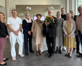 Eine Gruppe von neun Personen steht in einem modernen Empfangsbereich, einige halten Blumen. Sie scheinen an einer Feier oder Veranstaltung teilzunehmen.