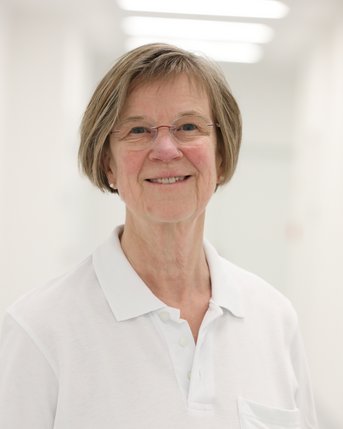 Eine ältere Frau mit Brille und kurzen Haaren trägt ein weißes Polohemd mit einer Brusttasche. Sie lächelt freundlich in einem hellen Flur.