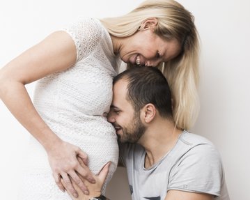 Ein Paar steht zusammen, die Frau trägt ein weißes Kleid und hält ihren Babybauch, während der Mann liebevoll auf den Bauch schaut.