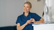 Eine lächelnde Frau in blauer Arbeitskleidung steht an einem Empfangstresen und hält einen Stift in der Hand.