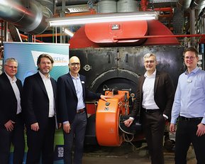 Fünf Personen stehen vor einem großen Kessel in einer Industrieumgebung, während sie an einem orangefarbenen Gerät arbeiten.