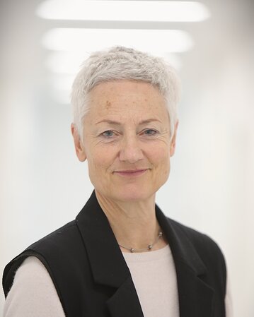 Eine Frau mit kurzem, grauem Haar trägt einen schwarzen Blazer über einem hellen Oberteil und lächelt in die Kamera.