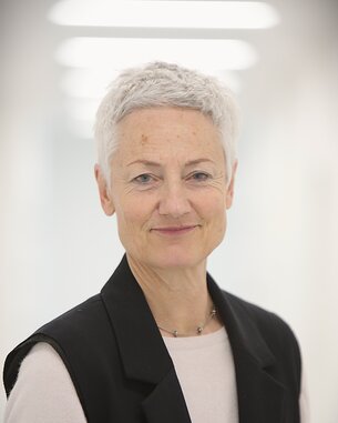 Eine Frau mit kurzem, grauem Haar trägt einen schwarzen Blazer über einem hellen Oberteil und lächelt in die Kamera.