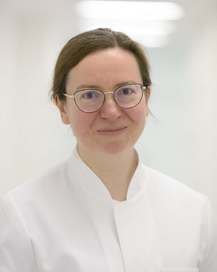Eine Person mit Brille und einem weißen Kittel lächelt freundlich in einem hellen, neutralen Raum.