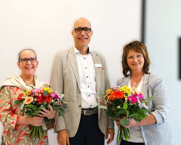 Drei Personen stehen zusammen und halten bunte Blumensträuße. Sie lächeln und tragen formelle Kleidung.