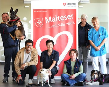 Eine Gruppe von sechs Personen und drei Hunden posiert vor einem Malteser-Logo. Die Personen tragen verschiedene Arbeitskleidung und lächeln.