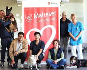 Eine Gruppe von sechs Personen und drei Hunden posiert vor einem Malteser-Logo. Die Personen tragen verschiedene Arbeitskleidung und lächeln.