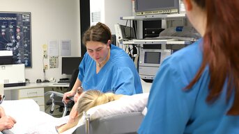 Vier medizinische Fachkräfte in blauen Kitteln kümmern sich um eine Patientin im Krankenhaus, während sie an einem Bett steht.