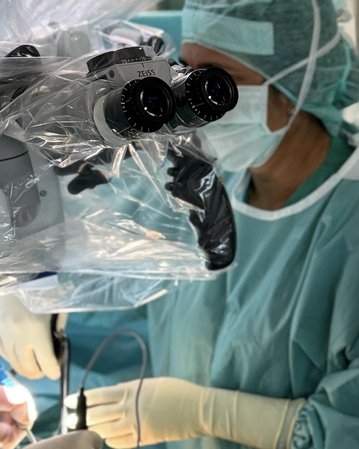 Ein Chirurg in einem Operationssaal trägt eine Schutzmaske und eine Haube, während er mit einem Mikroskop und chirurgischen Instrumenten arbeitet. Eine zweite Person assistiert ihm, während sie ebenfalls in OP-Kleidung gekleidet ist.