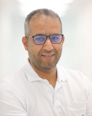 Ein Mann mit Brille und kurzem, grauem Haar lächelt freundlich. Er trägt ein weißes Polohemd mit einer Brusttasche. Der Hintergrund ist hell und unscharf.