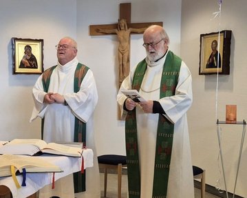 Zwei Priester in liturgischer Kleidung stehen in einer Kapelle, umgeben von religiösen Bildern und einem Kreuz. Ein Tisch mit einem offenen Buch ist im Vordergrund.