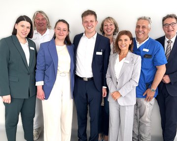 Eine Gruppe von acht Personen in formeller Kleidung, die zusammen lächeln. Sie stehen vor einem neutralen Hintergrund.