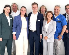 Eine Gruppe von acht Personen in formeller Kleidung, die zusammen lächeln. Sie stehen vor einem neutralen Hintergrund.