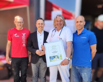 Vier Männer stehen zusammen, einer hält ein Zertifikat. Sie tragen unterschiedliche Kleidung, darunter ein rotes und ein blaues Shirt.