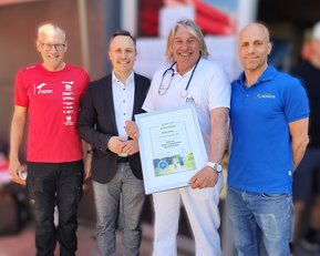 Vier Männer stehen zusammen, einer hält ein Zertifikat. Sie tragen unterschiedliche Kleidung, darunter ein rotes und ein blaues Shirt.