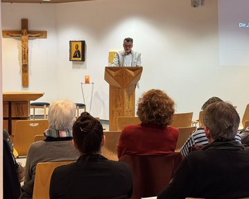 Ein Mann steht an einer Holzpult und spricht vor einer Gruppe von Zuhörern in einem Raum mit religiösen Symbolen.