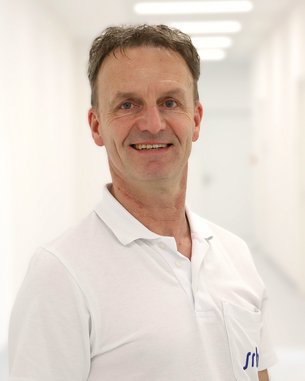 Ein Mann mit kurzen, grauen Haaren trägt ein weißes Polo-Shirt und lächelt in einem hellen Flur.