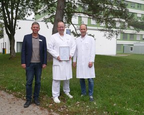 Drei Männer stehen im Freien, zwei tragen weiße Laborkittel und einer einen Anzug. Der Mann in der Mitte hält ein Dokument in der Hand. Im Hintergrund sind Gebäude und Bäume sichtbar.
