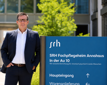 Mann in Anzug steht vor einem Gebäude mit einem Schild, das "SRH Fachpflegeheim Annahaus" und Informationen zum Haupt- und Warenanlieferungseingang zeigt.