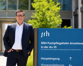 Mann in Anzug steht vor einem Gebäude mit einem Schild, das "SRH Fachpflegeheim Annahaus" und Informationen zum Haupt- und Warenanlieferungseingang zeigt.