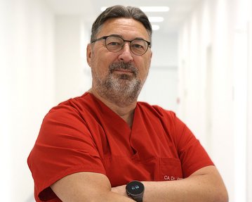 Ein Arzt in rotem Kittel steht mit verschränkten Armen in einem hellen Flur. Er trägt eine Brille und eine Smartwatch.