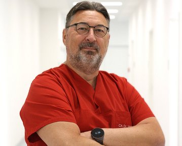 Ein Arzt in rotem Kittel steht mit verschränkten Armen in einem hellen Flur. Er trägt eine Brille und eine Smartwatch.