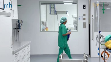 Eine Person in grüner OP-Kleidung und Haube geht durch einen modernen Krankenhausflur. Im Hintergrund sind medizinische Geräte sichtbar.