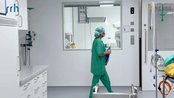 Eine Person in grüner OP-Kleidung und Haube geht durch einen modernen Krankenhausflur. Im Hintergrund sind medizinische Geräte sichtbar.