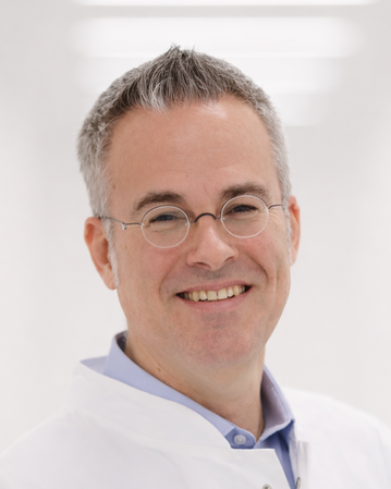 Ein lächelnder Mann in einem weißen Laborkittel steht vor einem hellen Hintergrund. Er trägt eine Brille und ein blaues Hemd.