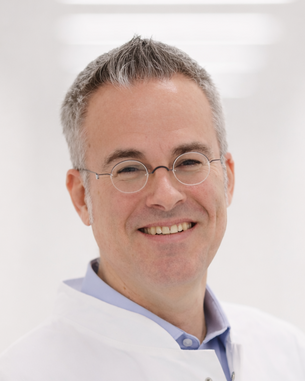 Ein lächelnder Mann in einem weißen Laborkittel steht vor einem hellen Hintergrund. Er trägt eine Brille und ein blaues Hemd.