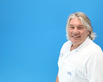 Ein Mann mit grauen Haaren trägt ein weißes Hemd und lächelt vor einem blauen Hintergrund.