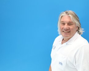 Ein Mann mit grauen Haaren trägt ein weißes Hemd und lächelt vor einem blauen Hintergrund.
