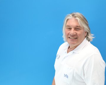 Ein Mann mit grauen Haaren trägt ein weißes Hemd und lächelt vor einem blauen Hintergrund.
