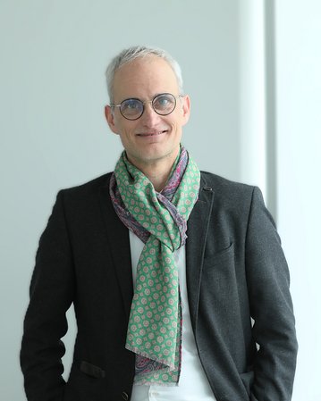 Mann mit Brille und grauen Haaren trägt einen bunten Schal und einen dunklen Blazer. Er steht in einem hellen Raum.