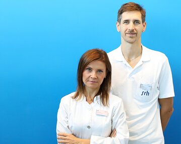 Zwei Fachkräfte in weißen Kitteln stehen vor einem blauen Hintergrund. Die Frau hat lange, braune Haare und der Mann hat kurzes, braunes Haar.