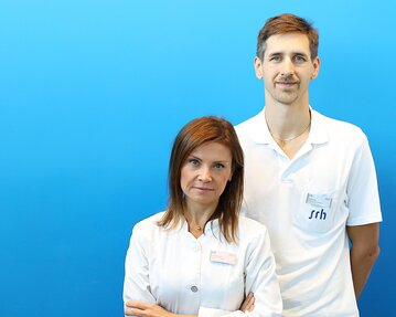 Zwei Fachkräfte in weißen Kitteln stehen vor einem blauen Hintergrund. Die Frau hat lange, braune Haare und der Mann hat kurzes, braunes Haar.